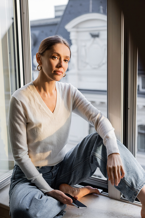barefoot woman in white jumper sitting on windowsill and looking at camera