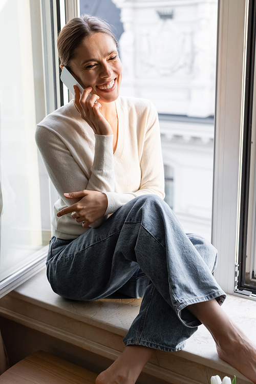 joyful woman sitting on windowsill during conversation on mobile phone