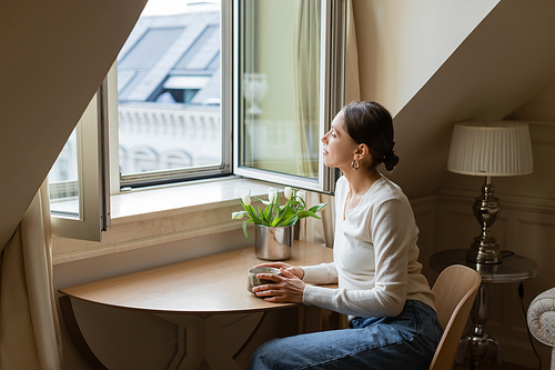 pensive woman sitting with clay cup near tulips and looking away through window