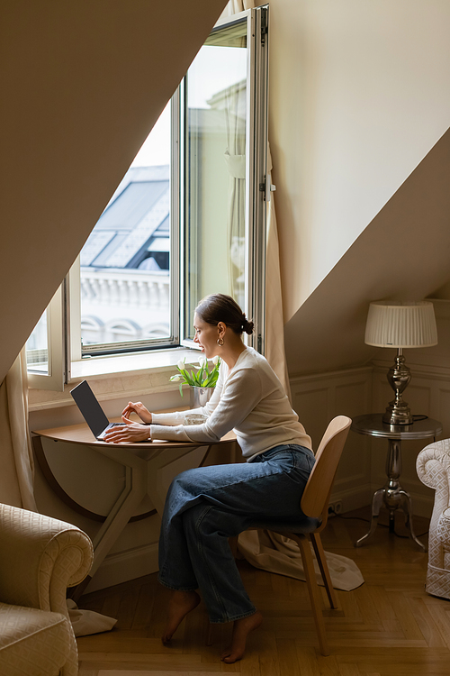 side view of woman typing on laptop in cozy attic room near window