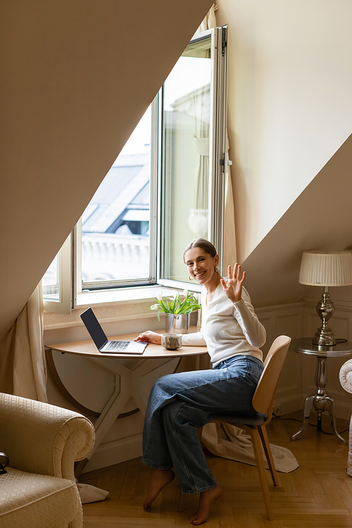 happy woman waving hand while sitting with laptop and clay cup near window