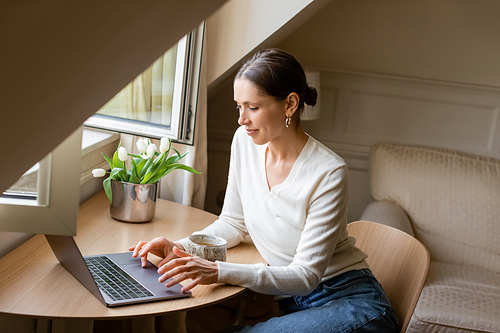 positive woman typing on laptop near window, clay cup and fresh tulips