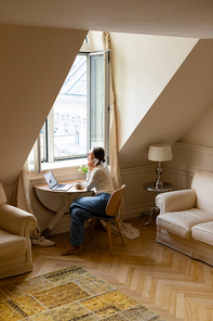 barefoot woman talking on mobile phone near laptop and window in cozy attic room
