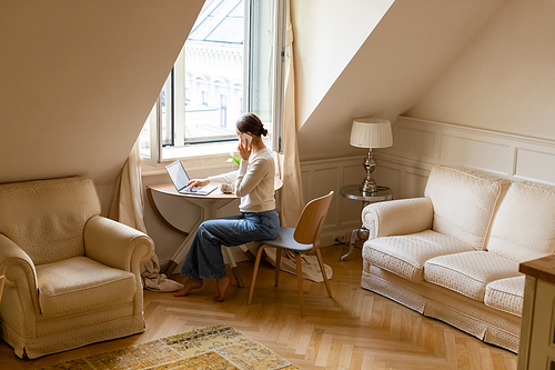 full length view of barefoot woman talking on smartphone near laptop in attic room