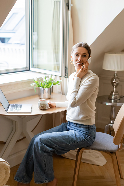 woman smiling at camera while talking on smartphone near laptop and tulips