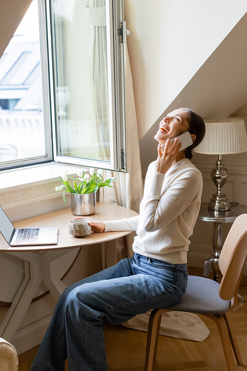 excited woman laughing during conversation on smartphone near clay cup, laptop and tulips