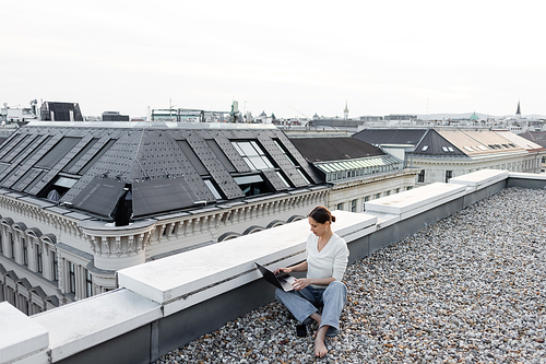 full length of barefoot woman in jeans working with laptop on rooftop of urban building