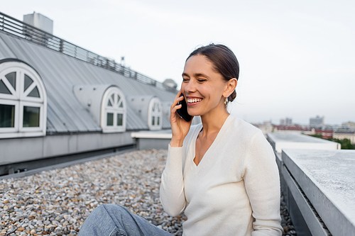 cheerful woman with closed eyes talking on mobile phone on rooftop of city building