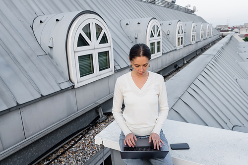 woman in white jumper typing on laptop near smartphone with blank screen on rooftop