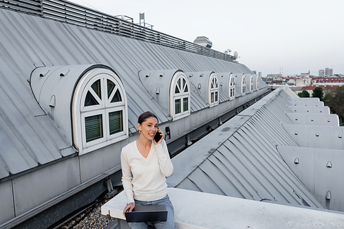 smiling woman with laptop talking on mobile phone while sitting on rooftop of city building