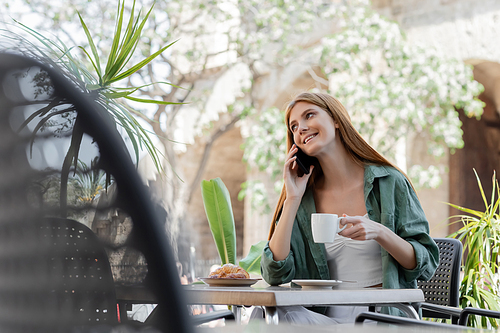 happy woman talking on smartphone and holding cup of coffee near croissant on table in cafe terrace