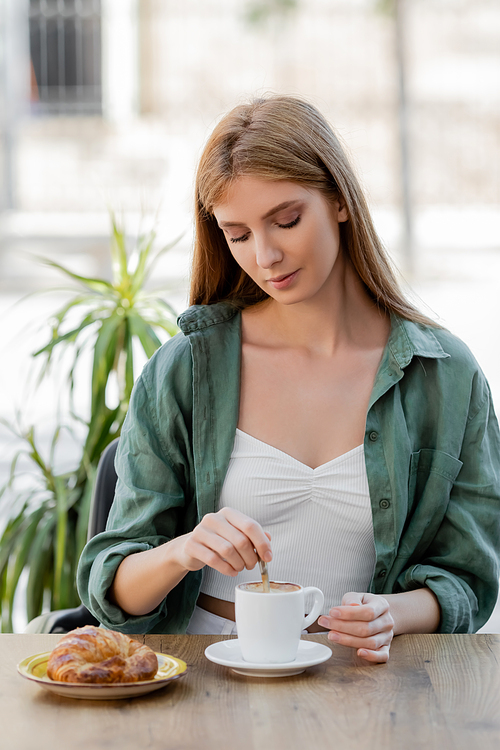 redhead woman stirring coffee with spoon near croissant on table in cafe terrace