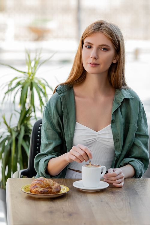 redhead woman stirring coffee with spoon and looking at camera on summer terrace