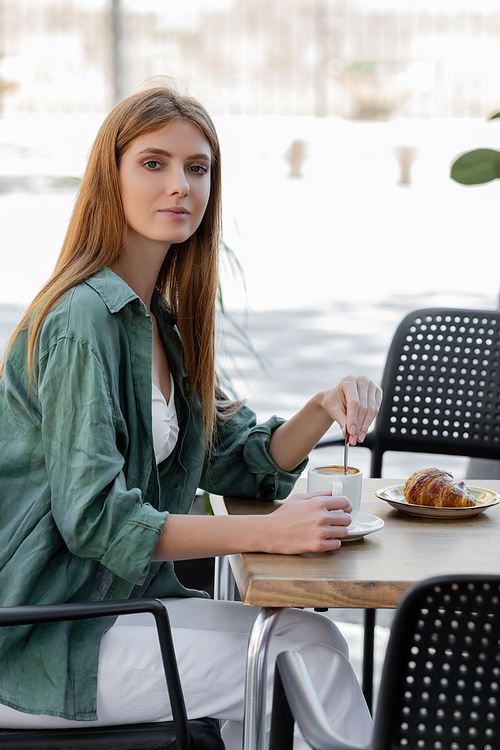 woman with red hair stirring coffee with spoon near tasty croissant in cafe terrace