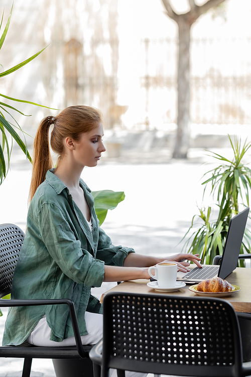 side view of woman with red hair using laptop near cup of coffee and tasty croissant in cafe terrace