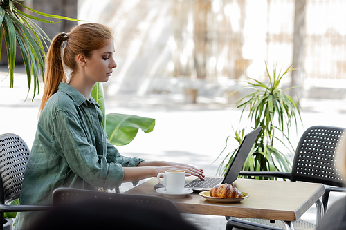 side view of freelancer with red hair using laptop near cup of coffee and tasty croissant in cafe terrace