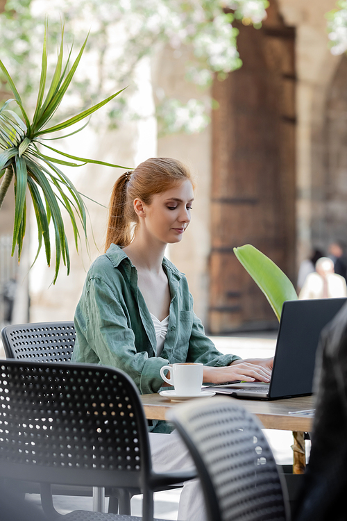 freelancer with red hair using laptop near cup of coffee in cafe terrace