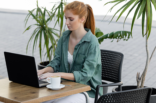 young woman with red hair using laptop near cup of coffee in cafe terrace