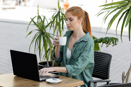 young woman with red hair using laptop and holding cup of coffee on summer terrace