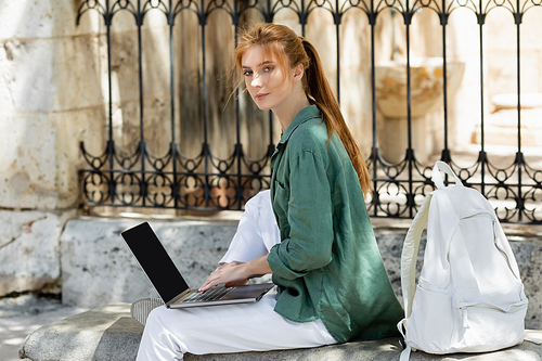 redhead freelancer sitting on concrete bench and using laptop near backpack