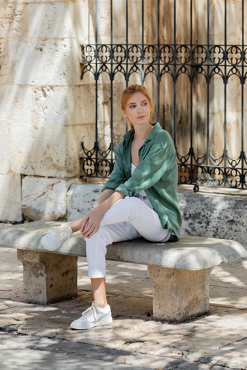 young redhead woman in green linen shirt sitting on concrete bench near forged fence in valencia