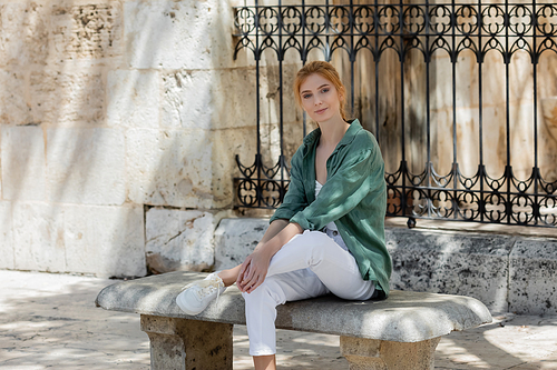 young woman with red hair in green linen shirt sitting on concrete bench near forged fence