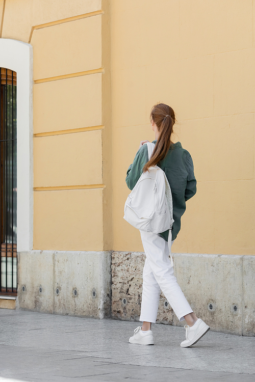 back view of redhead woman in green shirt and white pants holding backpack and walking near beige wall on street of valencia