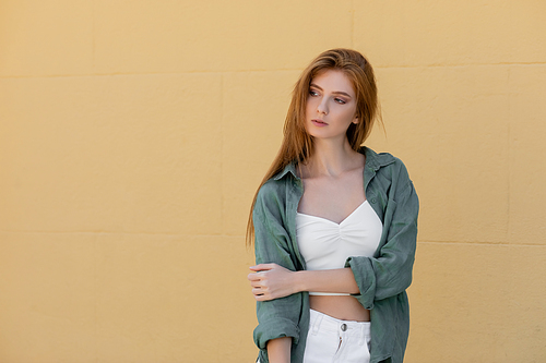 young redhead woman in green linen shirt posing near beige wall on street