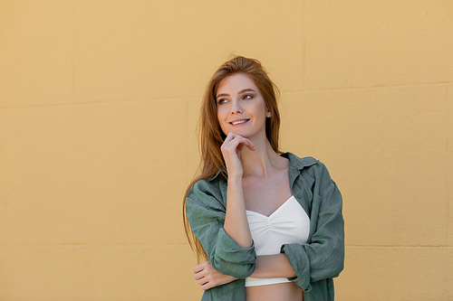 dreamy woman in green linen shirt smiling near beige wall on street