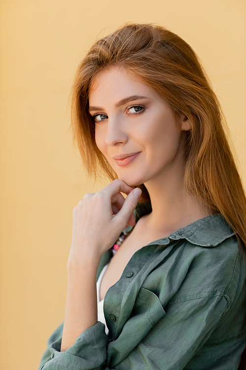 smiling and redhead woman in green linen shirt posing near beige wall