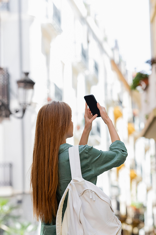 young redhead traveler with backpack taking photo on smartphone on urban street of valencia