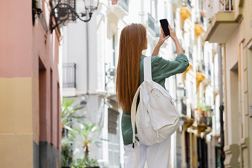 redhead woman with backpack taking photo on smartphone on urban street of valencia