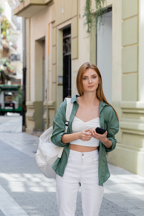 young redhead traveler with backpack holding smartphone on urban street of valencia