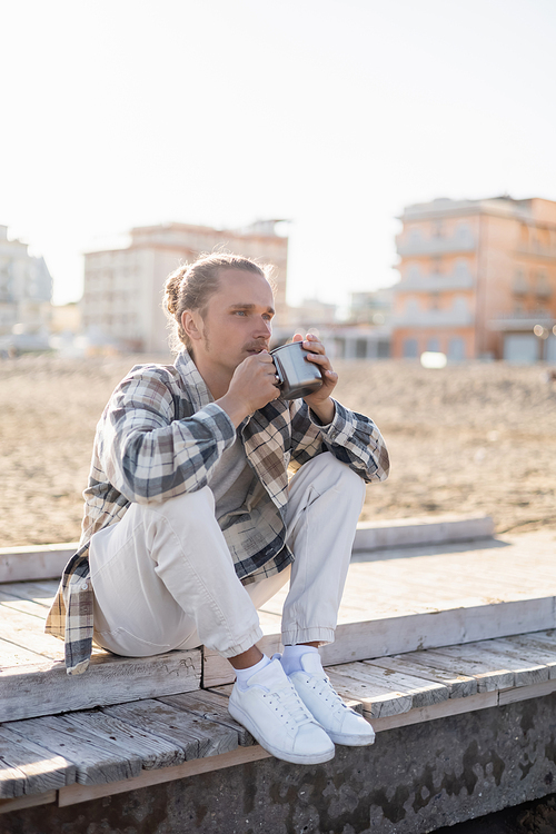 Long haired man holding cup while sitting on pier on beach in Italy