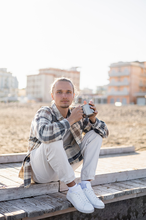 Young man in shirt holding metal cup while sitting on pier in Treviso