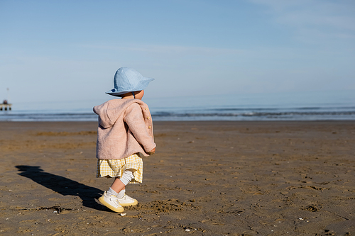 Baby girl in jacket and panama hat walking on beach in Italy