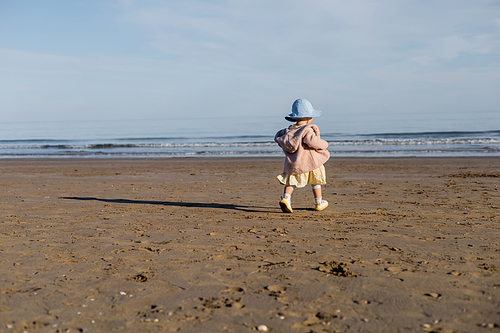 Back view of baby girl in panama hat walking on sandy beach in Italy