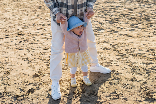 Man holding hands of cheerful baby in panama hat on beach