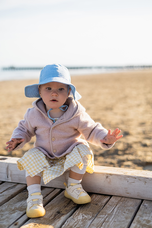 Baby girl sitting on wooden pier on blurred beach in Treviso