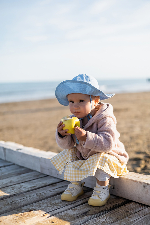 Child in panama hat holding fresh apple on pier on beach in Italy