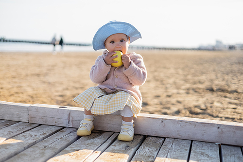 Baby girl in panama hat eating apple and looking at camera on wooden pier in Treviso