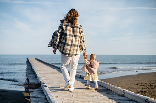 Back view of long haired man and toddler girl walking on wooden pier near adriatic sea in Italy