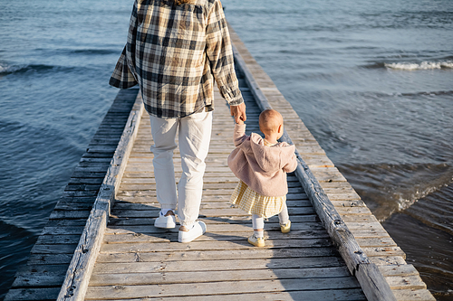 Back view of parent and baby girl walking on wooden pier above adriatic sea in Treviso