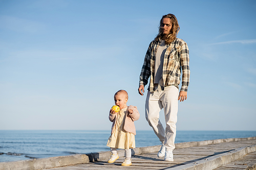 Young man walking near baby daughter with apple on pier on beach in Italy