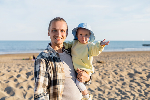 Smiling man looking at camera while holding baby daughter on beach in Treviso