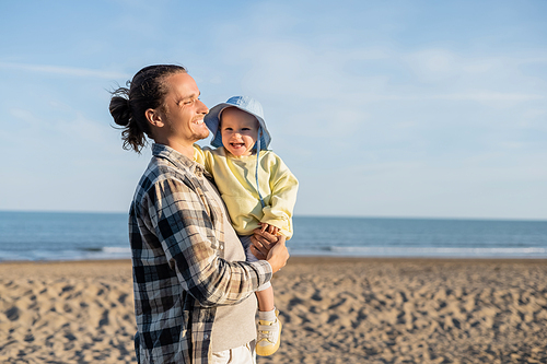 Long haired dad holding cheerful daughter on beach near sea in Italy