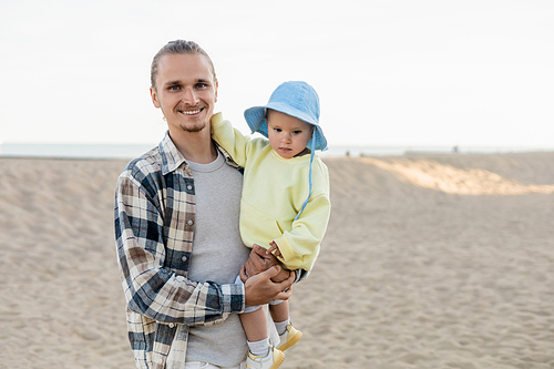 Positive man in shirt holding daughter and looking at camera on beach