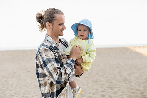 Long haired dad holding baby in panama hat on beach