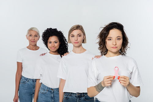 Tattooed woman holding ribbon of breast cancer awareness near interracial friends isolated on grey