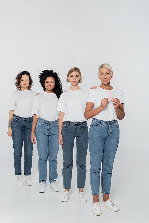 Senior woman holding ribbon of breast cancer awareness near multiethnic friends on grey background